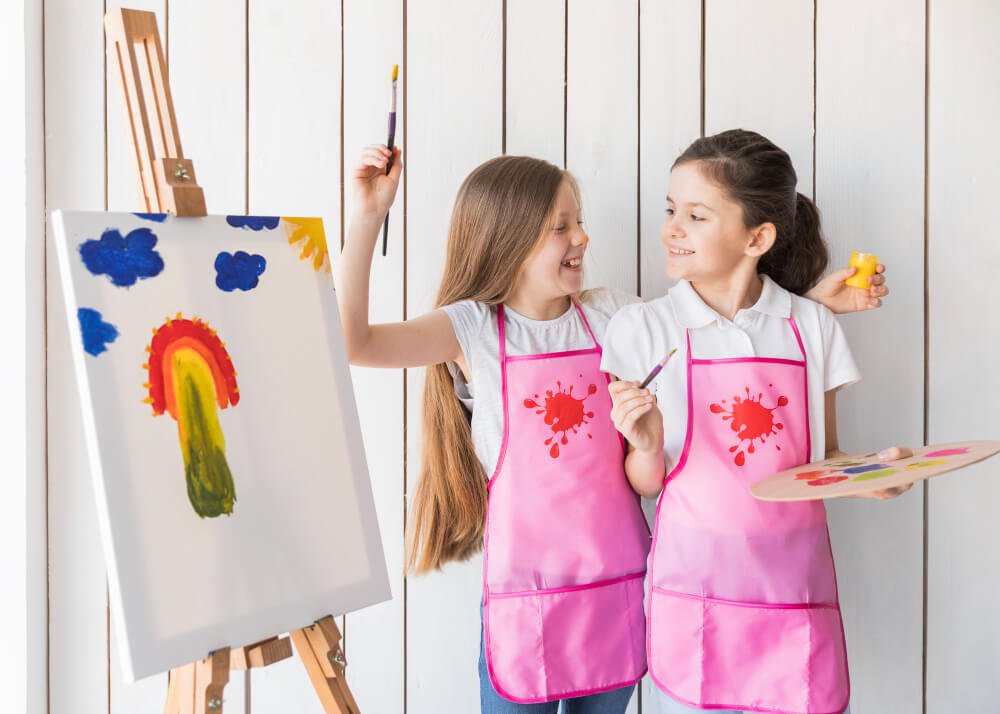 portrait-smiling-two-girls-pink-apron-making-fun-while-painting-canvas